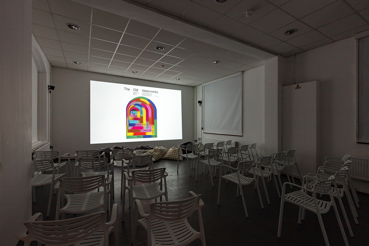 A large, darkened room with a projected image on the far wall. All walls and ceiling are white abd the floor is concrete. There are 4 rows of chairs and some bean bags at the front, below the projection.